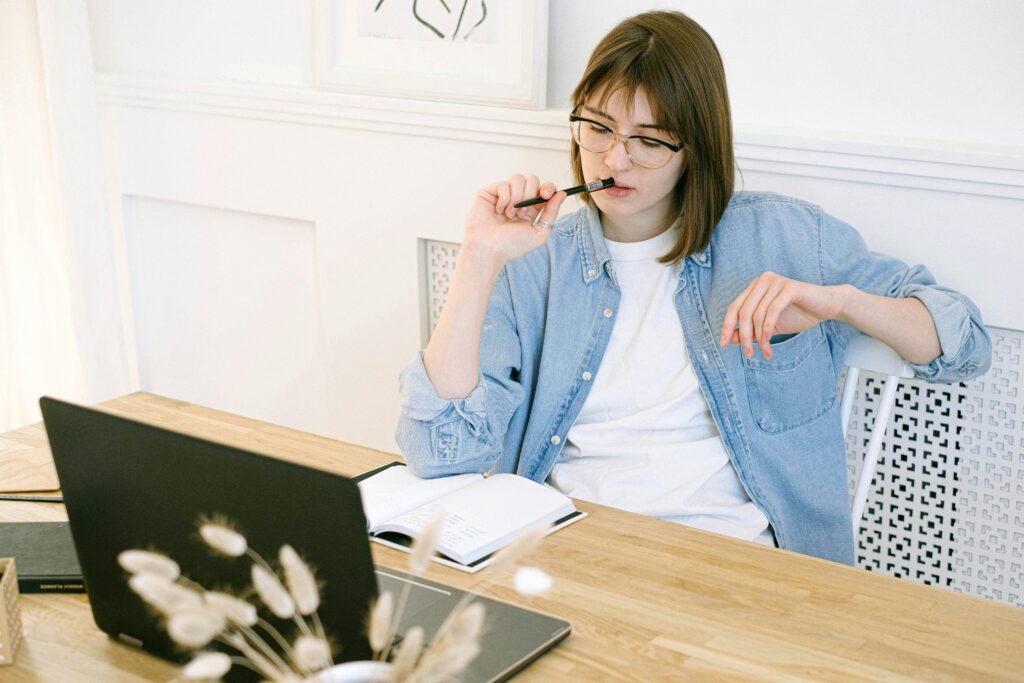 A lady sitting at a table with a notebook and a laptop computer in front of her. She is deep in thought as she looks down at her notebook.