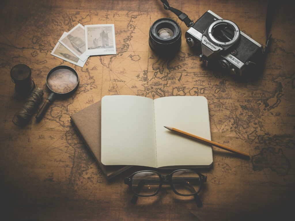 A notebook lies open, pages blank, waiting to be written in. Photographs, glasses and other items relating to a life lived lay around the book.